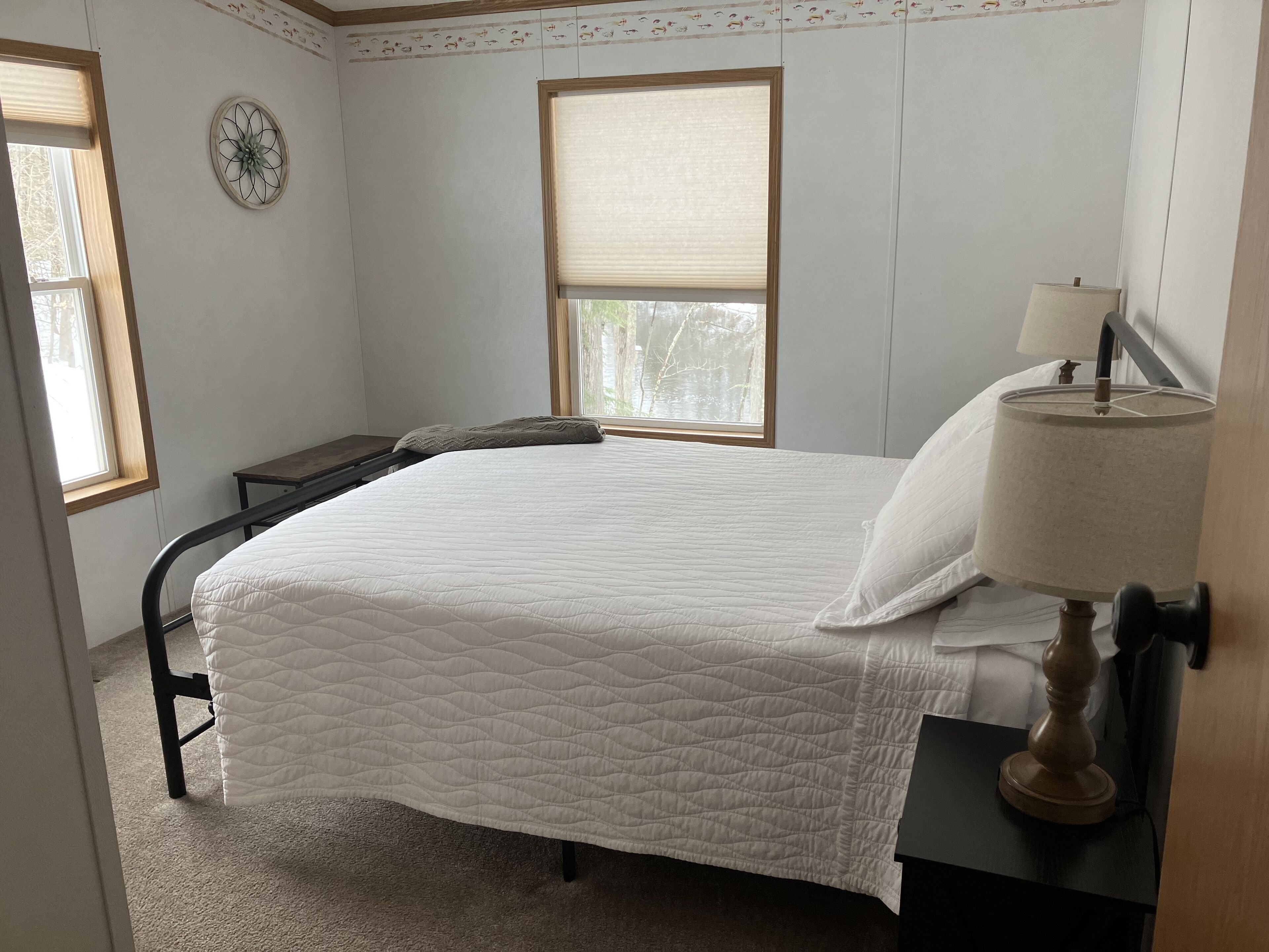 A bedroom with a bed, a nightstand, a lamp, a window, and a wall clock.