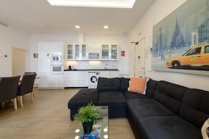 A modern living room with a black sectional sofa, a dining area with a table and chairs, a kitchen with white cabinets, a washing machine, and a painting of a cityscape on the wall. - 4br 2bth best location SOl, new (Madrid)