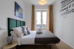 A neatly made bed with a green headboard, a bedside table with a lamp, and a window with curtains.