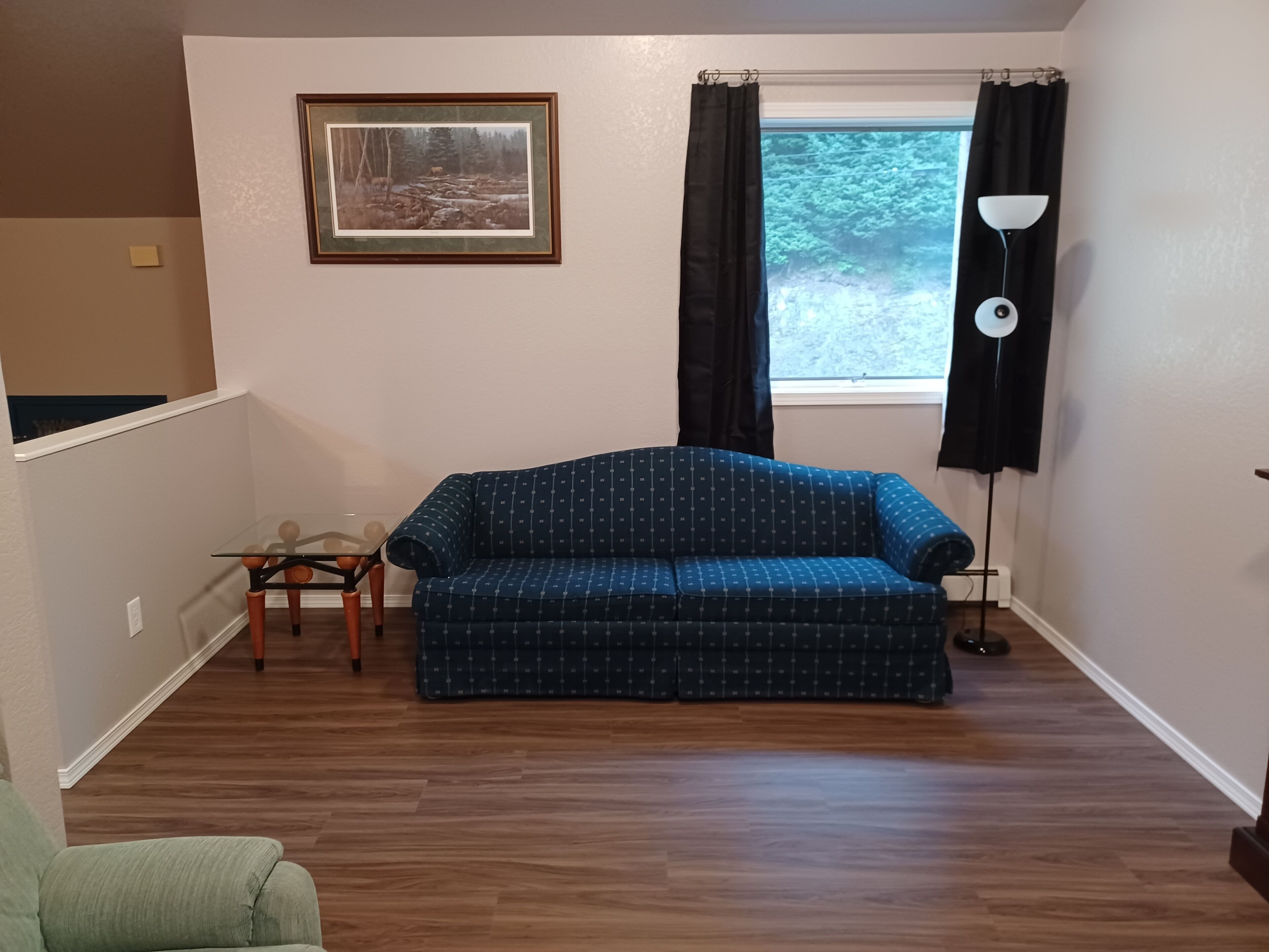 A living room with a blue patterned sofa, a small table with chairs, a framed picture on the wall, a window with curtains, and a floor lamp.