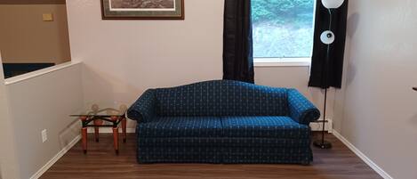 A living room with a blue patterned sofa, a small table with chairs, a framed picture on the wall, a window with curtains, and a floor lamp.