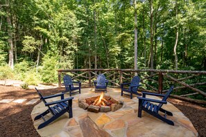 Un espace extérieur aménagé avec des chaises Adirondack et un foyer en pierre, le tout entouré d’une forêt.