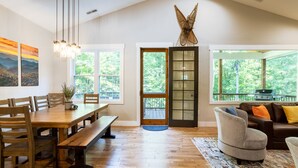 Un amplio comedor con una mesa de madera y bancos, ventanas grandes que ofrecen vistas a zonas verdes y una zona de estar acogedora con sofá y cojines.