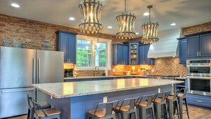 A modern kitchen with blue cabinets, a large island with bar stools, stainless steel appliances, and unique pendant lighting. - Brush Creek Pool House | Luxurious 10BR Home with Pool, Fire Pit, Game Room! (Fairview)
