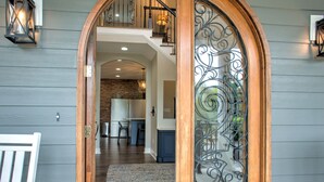 A wooden door with a decorative glass panel and a metal gate, leading to a staircase and a kitchen area with a brick wall and modern appliances. - Brush Creek Pool House | Luxurious 10BR Home with Pool, Fire Pit, Game Room! (Fairview)