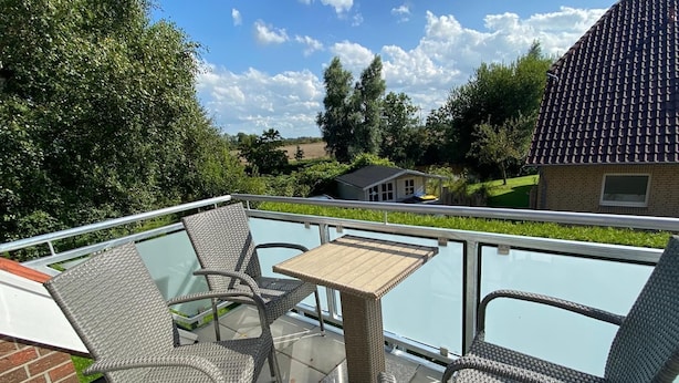 A balcony with wicker furniture and a glass table, overlooking a residential area with houses and greenery. - Ferienwohnung Am Harlebogen 10 Whg. 4 (Wittmund)