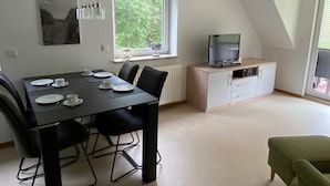 A dining area with a table set for four, a television on a cabinet, and a green armchair. - Ferienwohnung Am Harlebogen 10 Whg. 4 (Wittmund)
