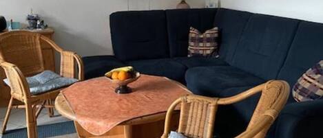 A living room with a dark blue sofa, two wicker chairs, a small round table with a fruit bowl, and a lamp.