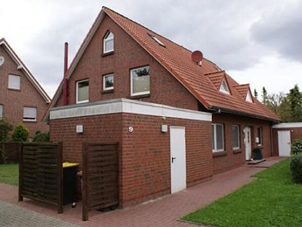 Une maison à deux étages, construite en briques, avec un toit recouvert de tuiles rouges, une porte d’entrée et un petit garage attenant.