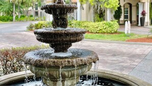 A multi-tiered stone fountain with water flowing from the top tier, set in a landscaped area with a building in the background. - Spacious & Cozy Home-Near all Disney Parks (Davenport)