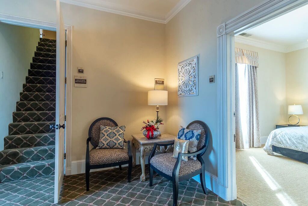 A hotel room with a staircase, two chairs, a small table with a lamp, and a bed.