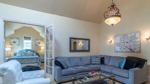 A living room with a grey sofa, a white armchair, a glass coffee table, and a chandelier. There is a bed in the adjacent room with a window, and a painting on the wall. - Penthouse King Suite•Private Floor•Monterey (Salinas)