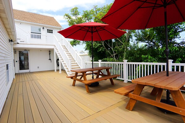 Una terraza de madera con mesas de picnic y sombrillas rojas, barandas blancas y una escalera que lleva a un segundo nivel.