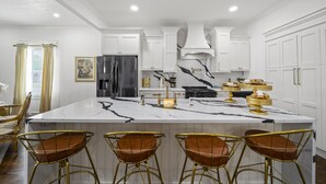 A modern kitchen with white cabinetry, a marble island, gold bar stools, and a stainless steel refrigerator.