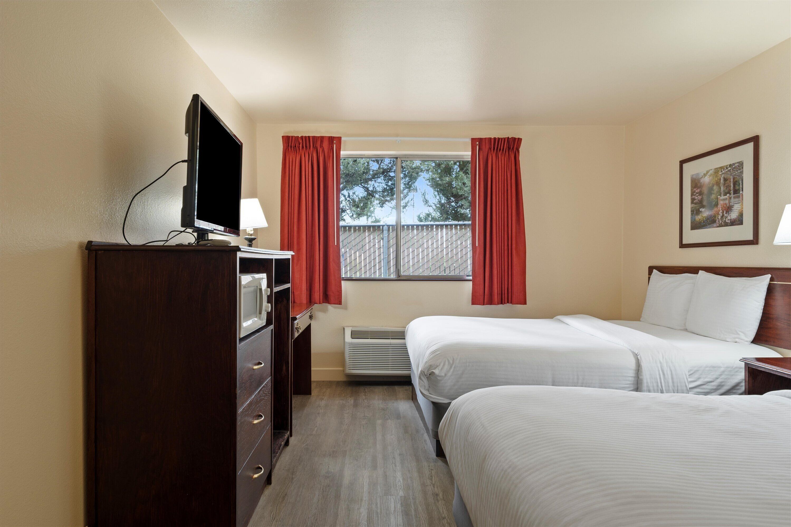 A hotel room with two beds, a TV, a desk, and a window with curtains.