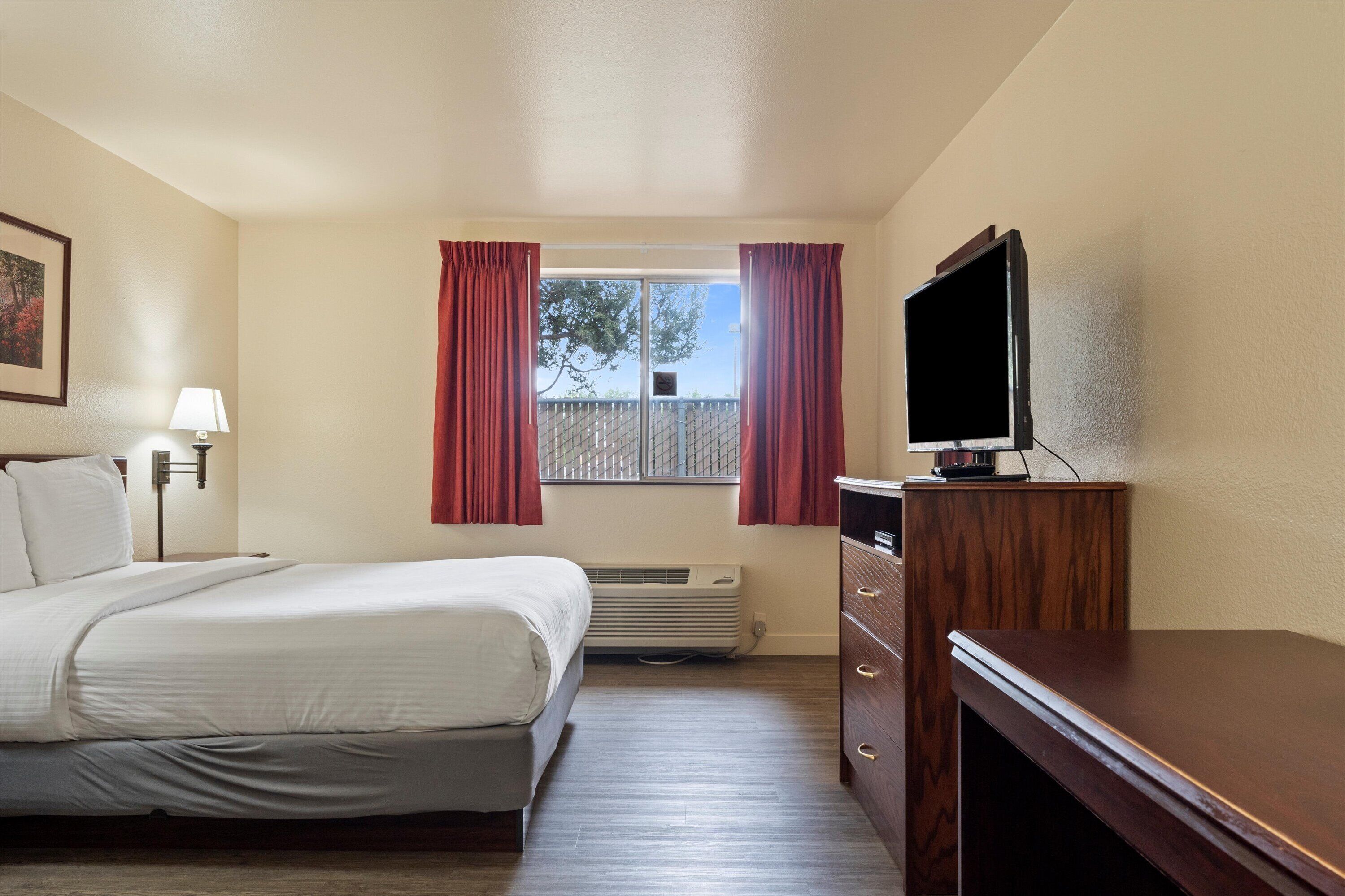 Une chambre d’hôtel avec un lit, un téléviseur posé sur une commode en bois, une fenêtre avec des rideaux, une lampe fixée au mur et un radiateur.
