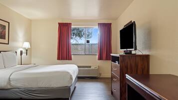 A hotel room with a bed, a TV on a wooden dresser, a window with curtains, a wall-mounted lamp, and a heater.