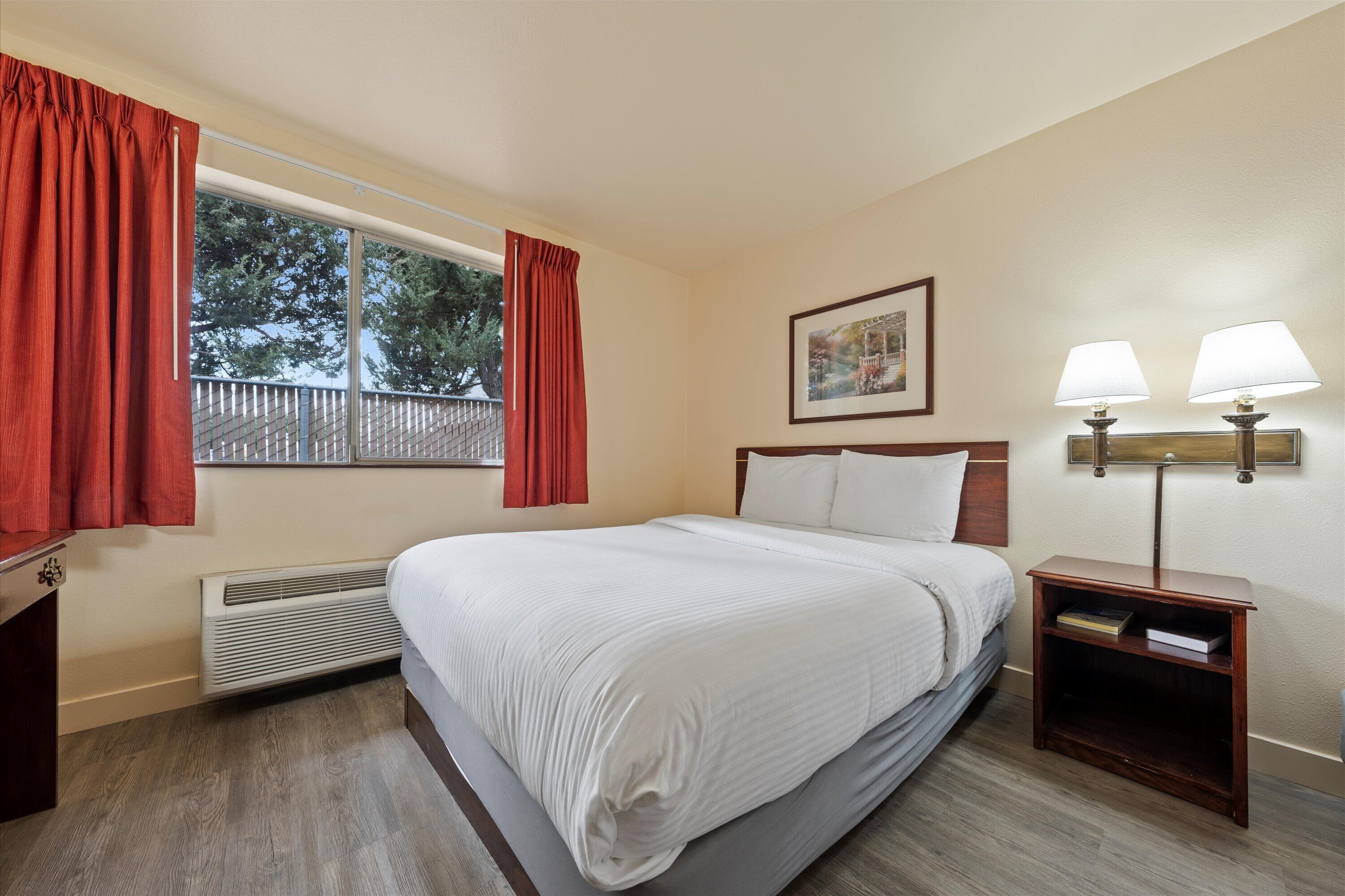 Habitació d'hotel amb un llit gran, una finestra amb cortines vermelles, un radiador, una tauleta de nit amb dues làmpades i un quadre encimbellat a la paret.