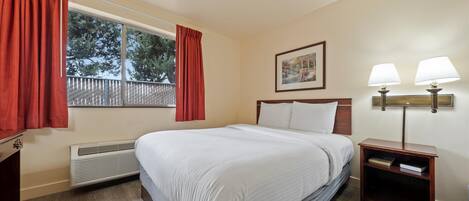 Habitació d'hotel amb un llit gran, una finestra amb cortines vermelles, un radiador, una tauleta de nit amb dues làmpades i un quadre encimbellat a la paret.