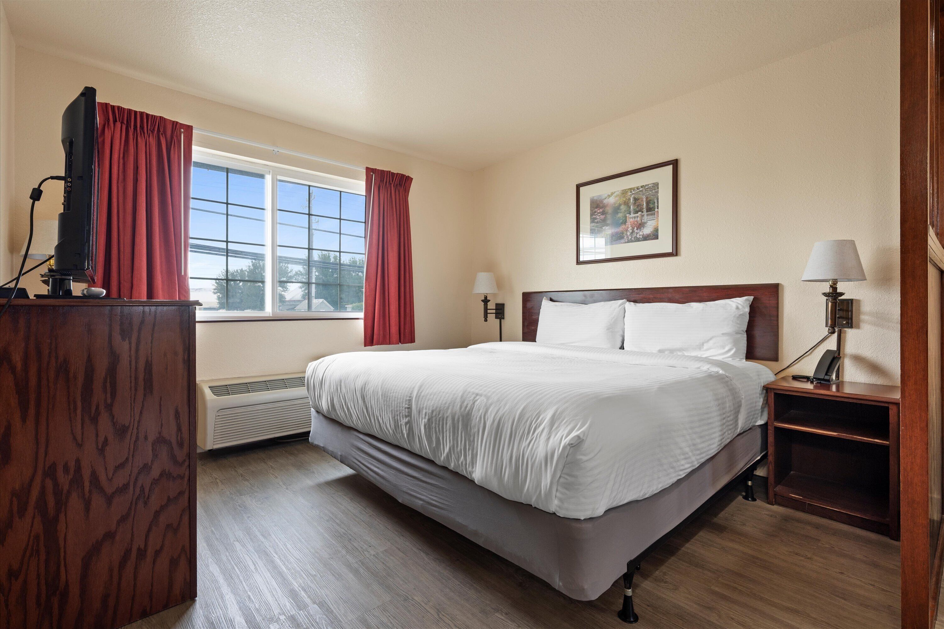 Una habitación de hotel con una cama grande, un escritorio con una computadora, una ventana con cortinas rojas y un cuadro en la pared.