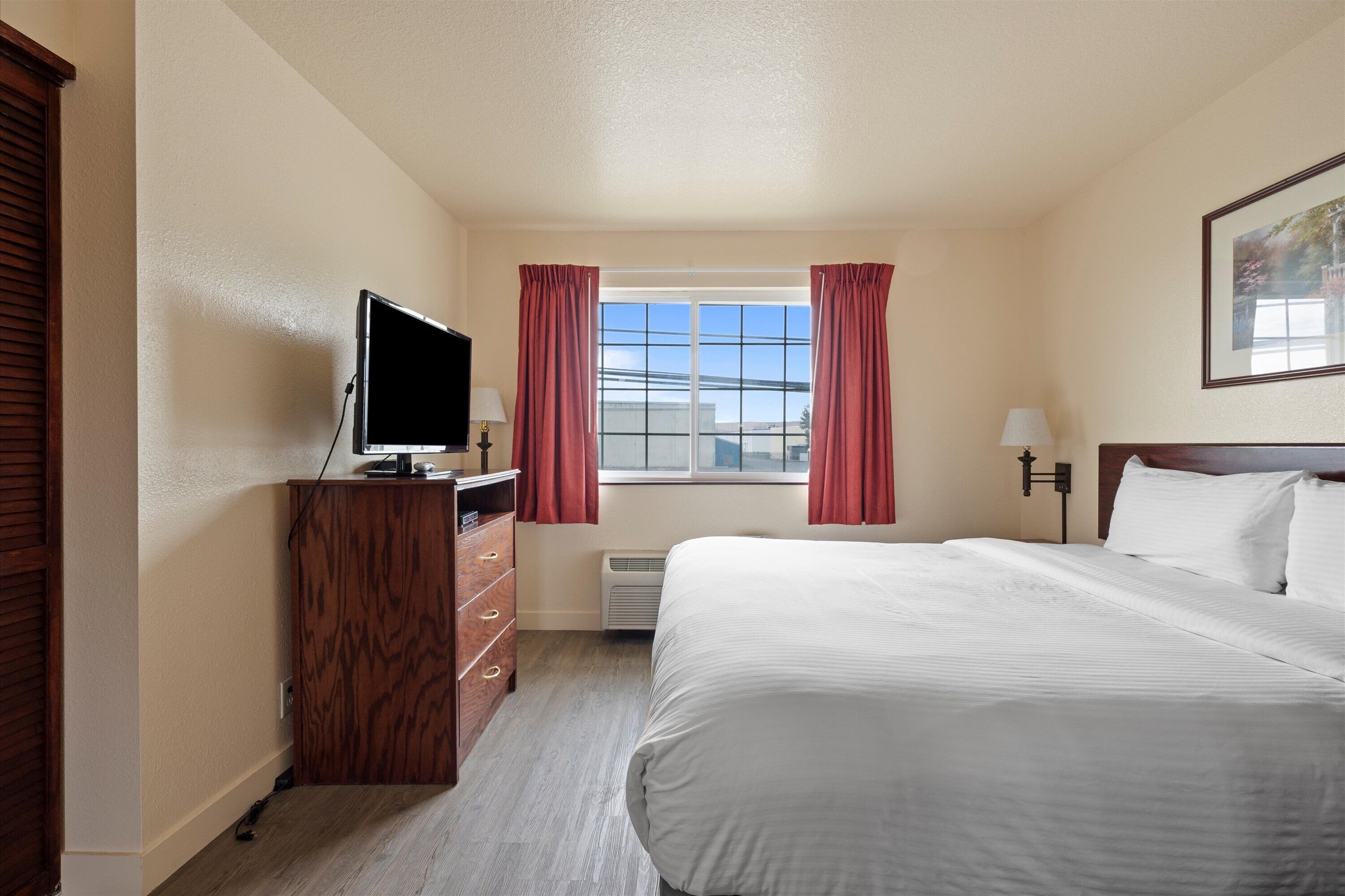 A hotel room with a bed, a TV on a wooden dresser, a window with curtains, and a wall-mounted lamp.