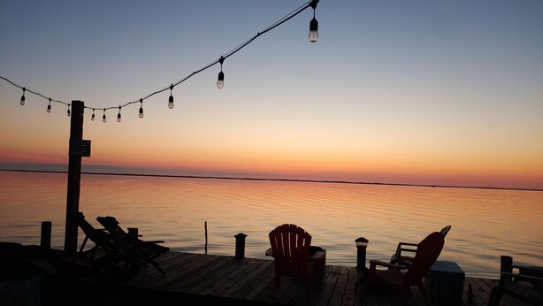 A wooden deck with chairs and a table, overlooking a calm body of water during sunset.