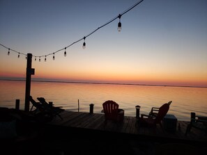 A wooden deck with chairs and a table, overlooking a calm body of water during sunset.