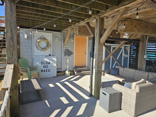 A covered outdoor area with a wooden ceiling, string lights, a 'SLOW NO WAKE' sign, a green chair, a wicker sofa, and a small table.
