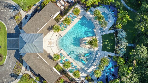 Eine Vogelperspektive zeigt ein Resort mit einem zentralen Schwimmbecken, Liegebereichen und Palmen.