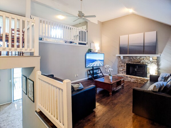 A spacious living room with a high ceiling, a fireplace, a television, and a balcony with a railing.