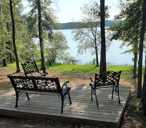 Una terraza de madera con dos bancos mirando a un lago, rodeada de árboles.