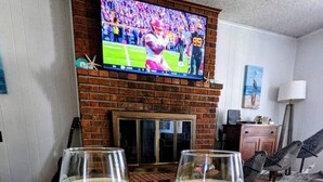 Una sala de estar con chimenea de ladrillo, un televisor mostrando un partido de fútbol y dos vasos de una bebida amarilla sobre una mesa de madera.