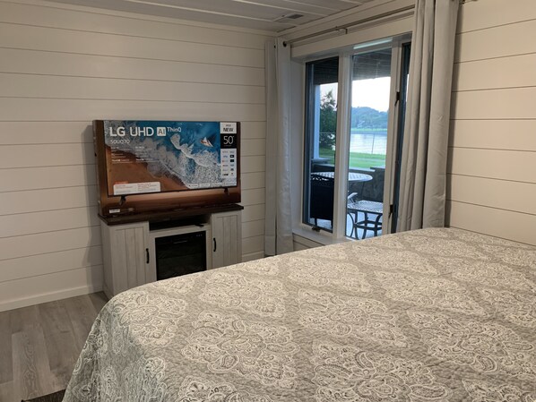 A bedroom with a bed, a television on a stand, a window with a view of a patio, and a wall-mounted thermostat.