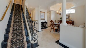 A grand staircase with ornate black and gold carpeting, leading to a dining area with a chandelier and a white column.
