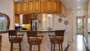 A kitchen with wooden cabinets, a stainless steel refrigerator, and a bar area with three stools.