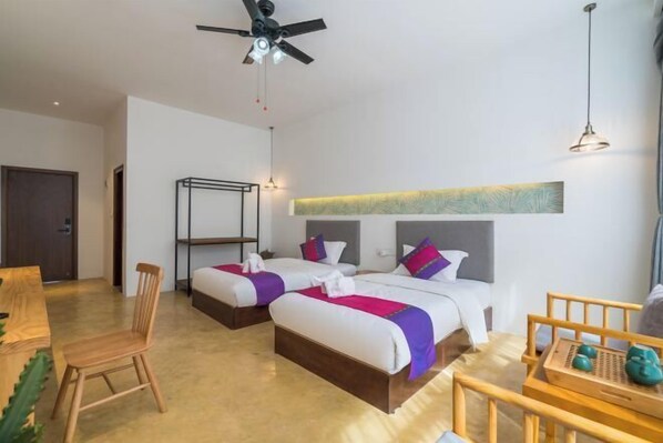Habitación de hotel con dos camas, ventilador de techo, una silla de madera, una mesita con bandeja y un estante en la pared.
