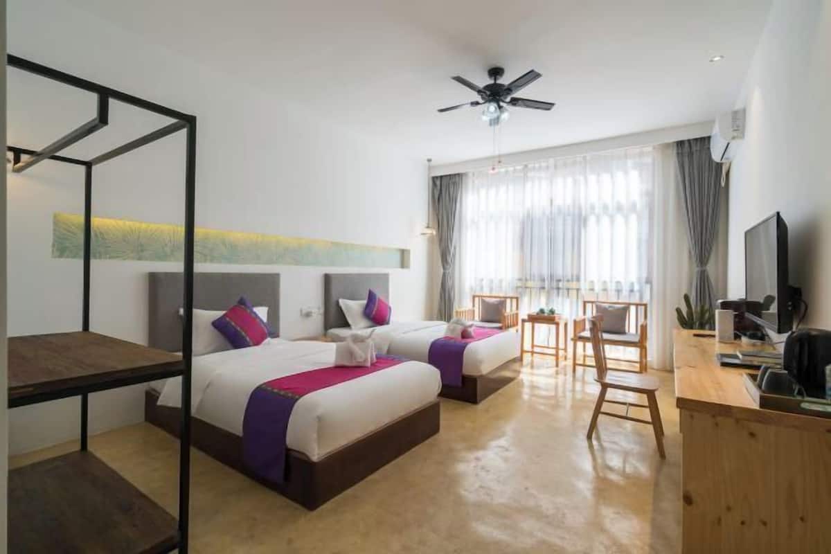 A hotel room with two beds, a desk, a chair, a TV, and a ceiling fan.
