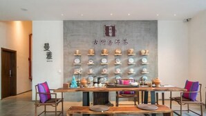 A dining area with a long wooden table, chairs, and benches. There are shelves with various items on the wall behind the table. - Manya Hotel (Menghai)