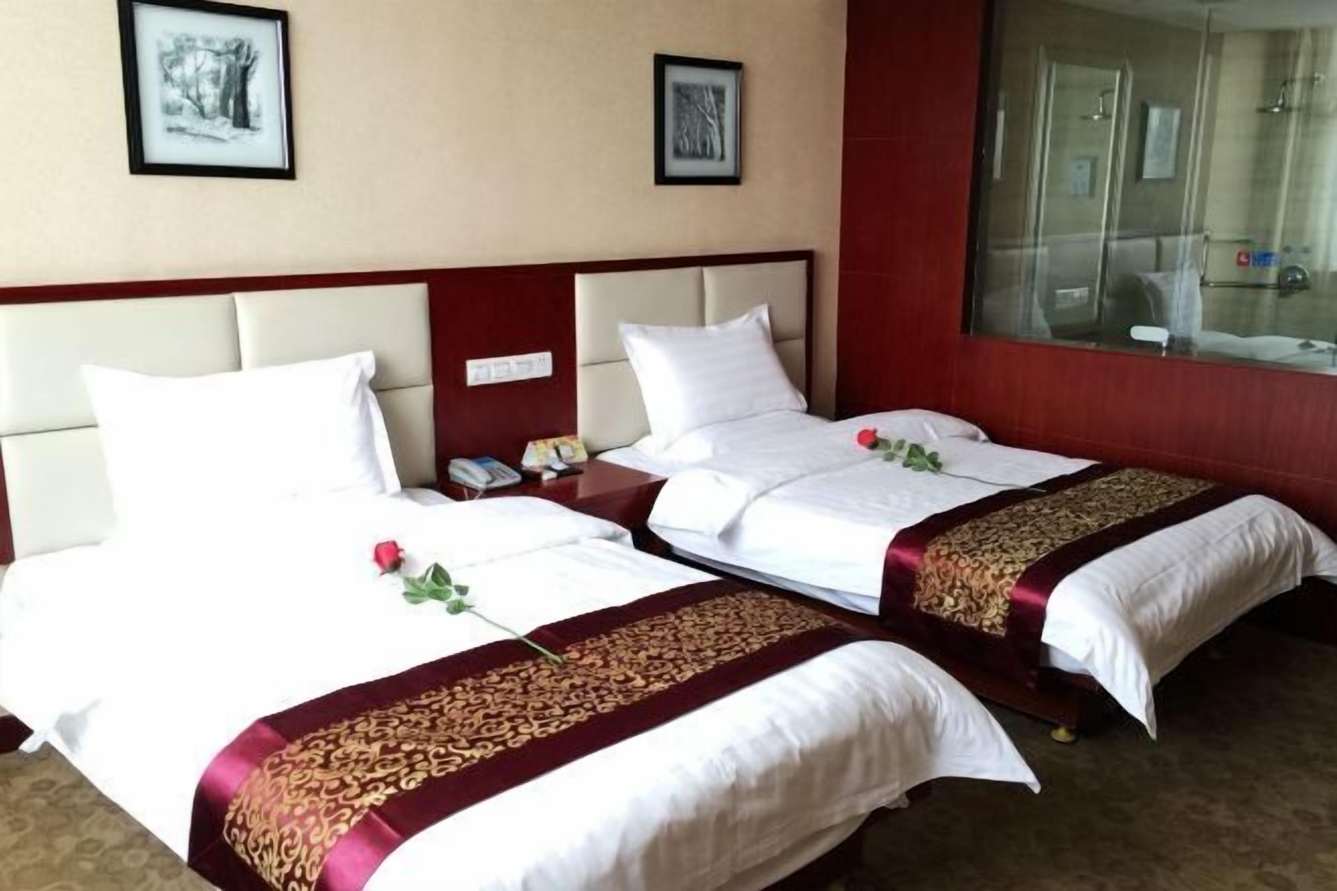 A hotel room with two beds, each with white linens and a red and gold decorative bedspread. There are framed pictures on the wall, a bedside table with a phone, and a large mirror.