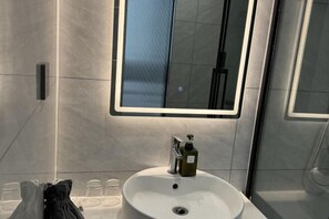 A modern bathroom with a large mirror, a white sink, and a glass shower enclosure. - Qi Ting Hotel (Linquan)