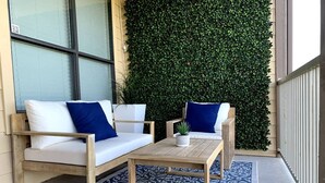 Une terrasse avec un canapé blanc et deux coussins bleus, une table en bois sur laquelle est posée une plante en pot, et un tapis à motifs.
