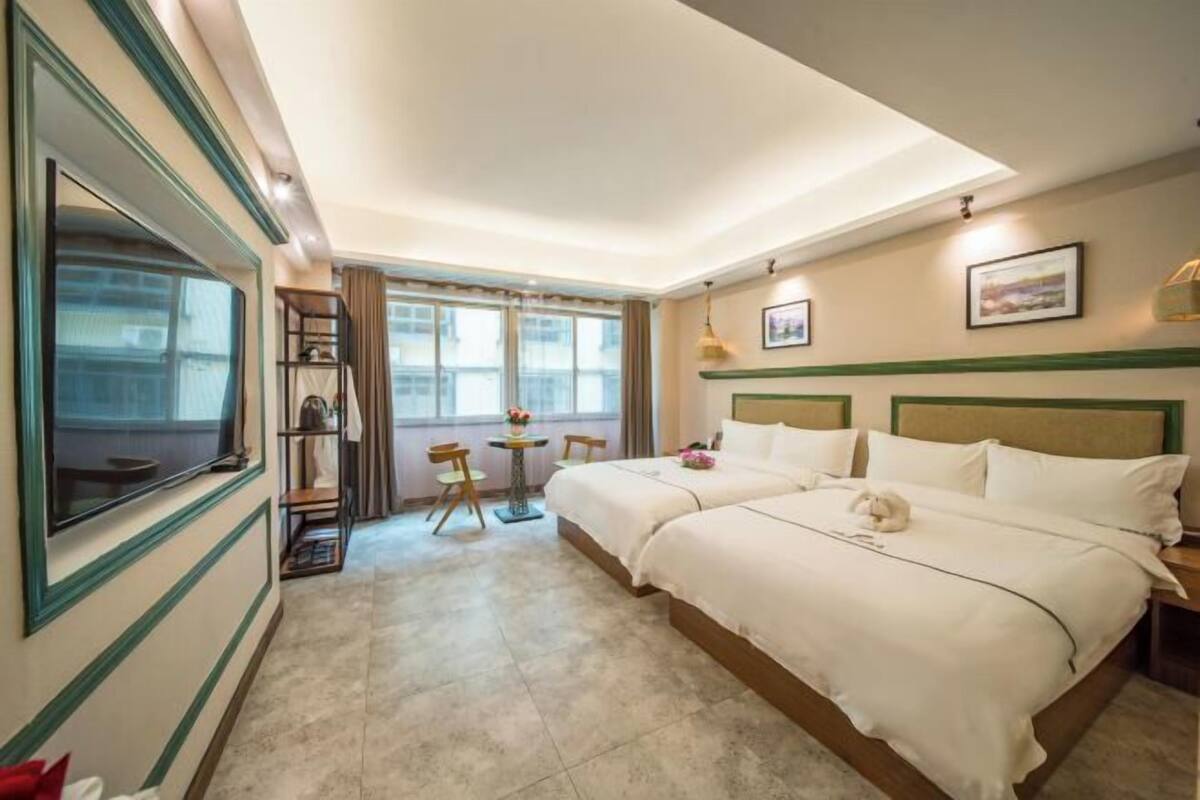 Quarto de hotel com duas camas, um espelho grande, uma mesinha com cadeiras e uma janela com cortinas.