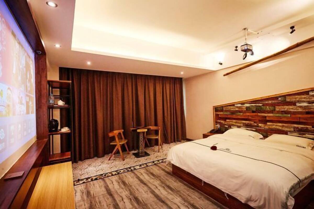 Quarto de hotel com cama, escrivaninha, duas cadeiras, televisão e cabeceira de madeira.