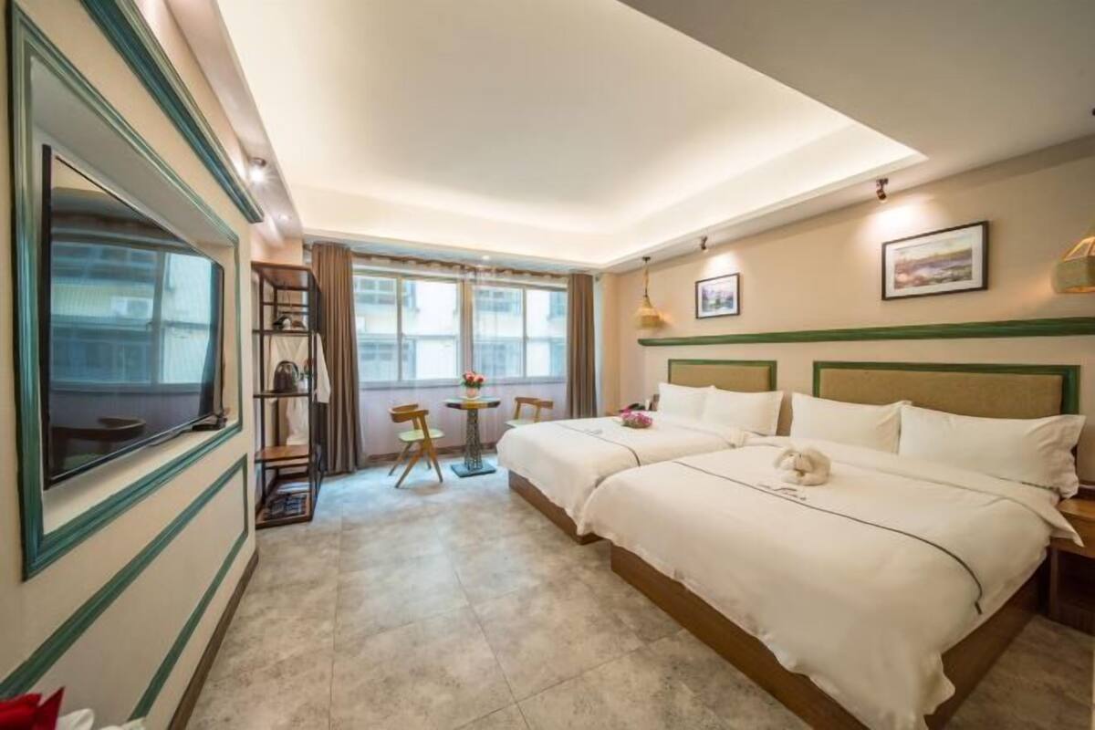 Quarto de hotel com duas camas, uma janela grande, uma mesa pequena com cadeiras e uma televisão.