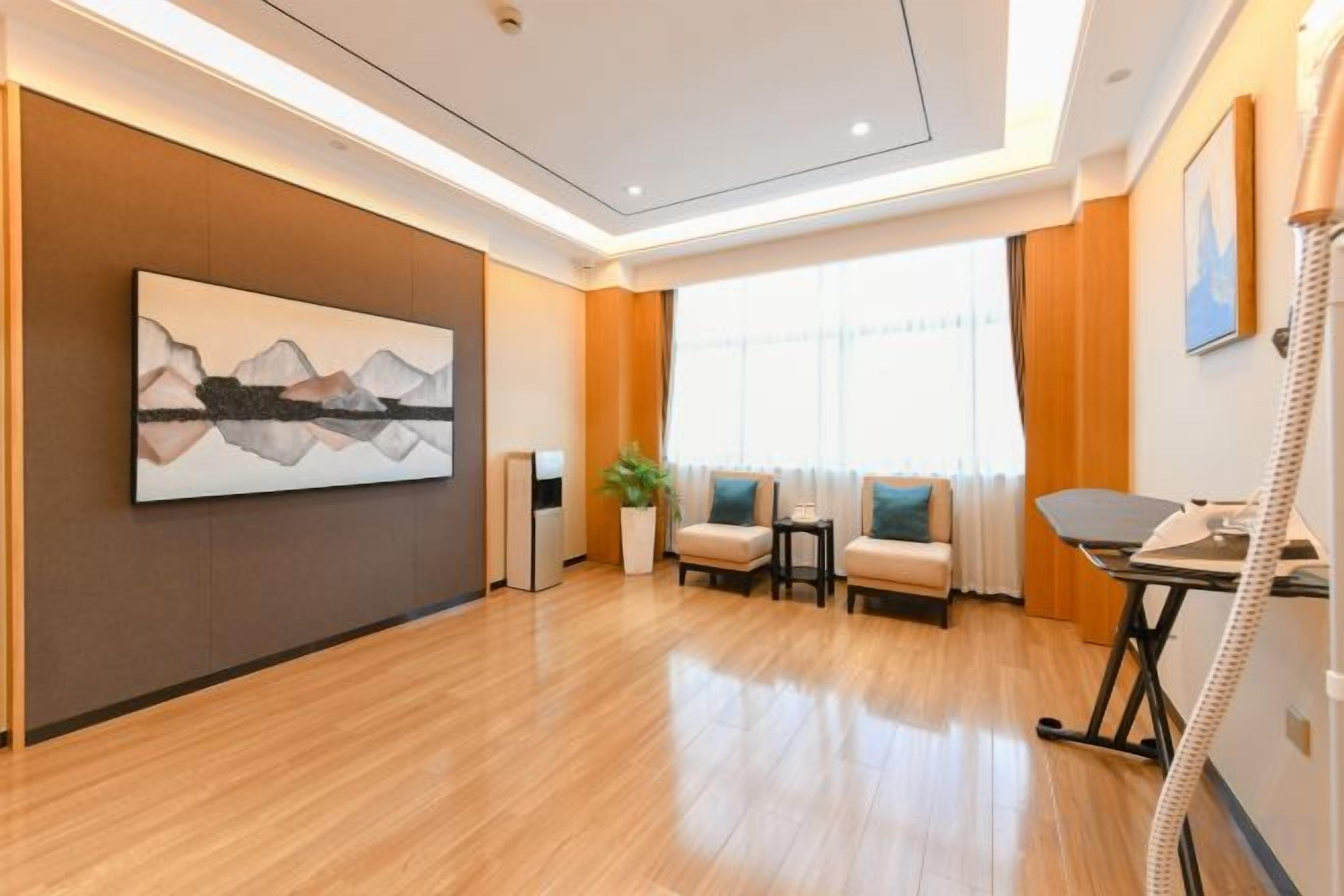 A hotel room with a wooden floor, a painting on the wall, a desk with a telephone, and a window with curtains.