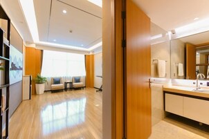 Una habitación de hotel moderna con suelo de madera, un ventanal grande, una zona de estar con sofás y un baño con un espejo grande y lavamanos.