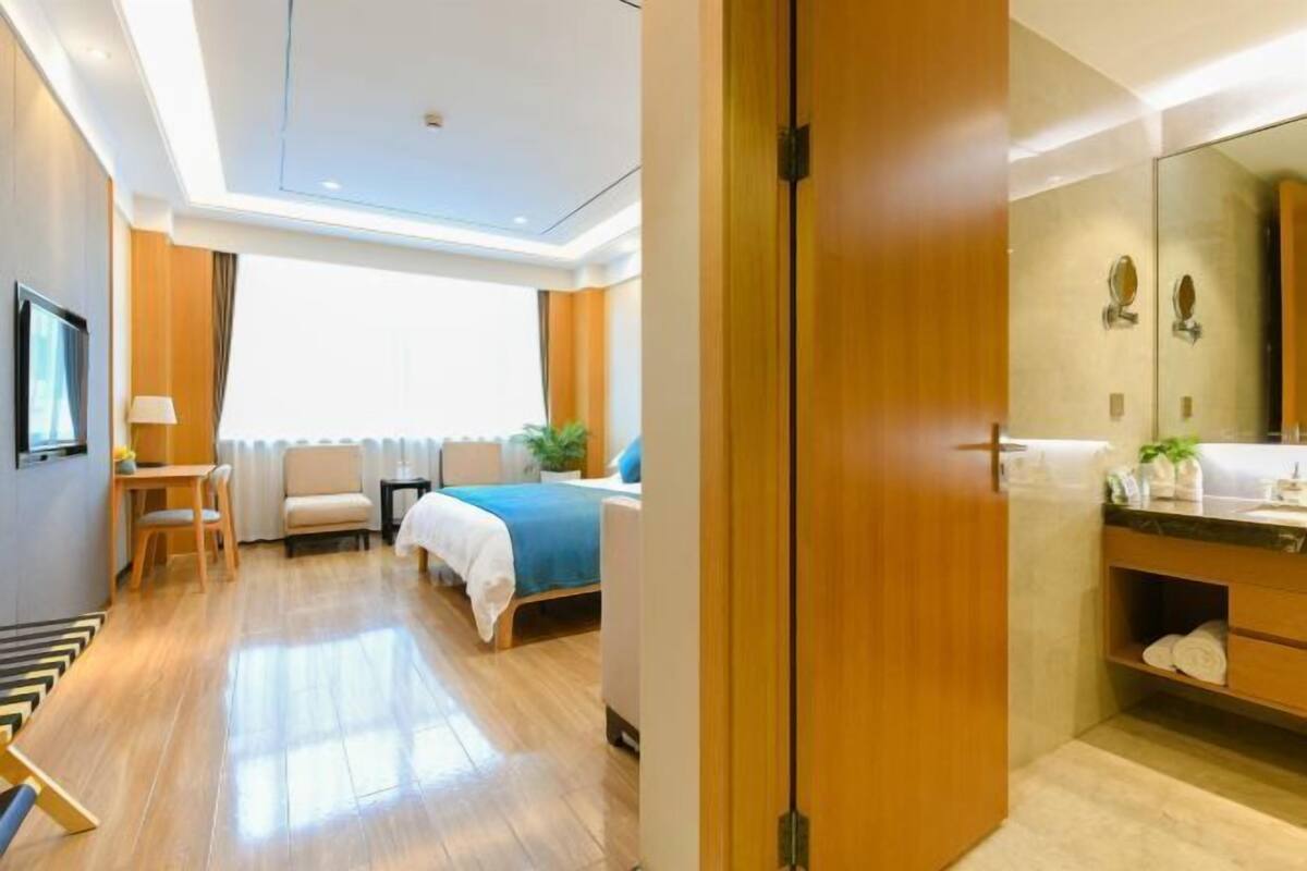 A hotel room with a bed, a desk, a chair, a window with curtains, a mirror, and a bathroom with a sink and towel rack.