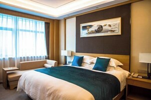 Habitación de hotel con una cama grande, una mesita de noche, una silla, un cuadro en la pared y una ventana con cortinas.