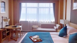 Ett hotellrum med en säng, en soffa, ett skrivbord med en stol, ett fönster med gardiner och en lampa.