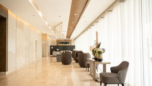 A modern hotel lobby with a clean, polished floor, a reception desk, comfortable seating, and large windows with sheer curtains.
